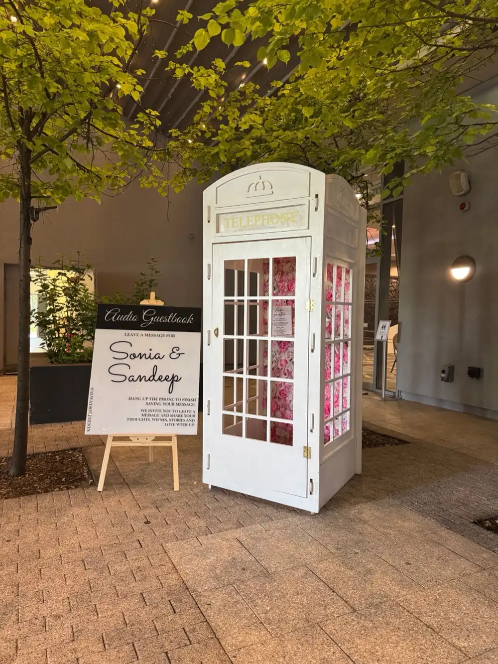 White telephone box audio guest book with a pink flower wall inside and a welcome sign on an easel beside it