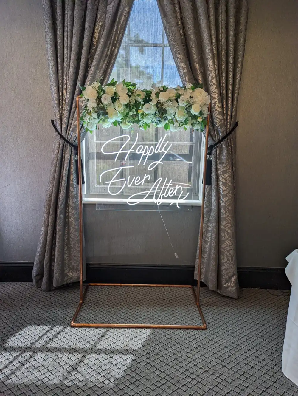 Neon Happily Ever After sign with a white floral garland on a copper stand beside draped curtains